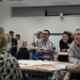 Groups of people are sat at a table and listening to a talk from a presenter.
