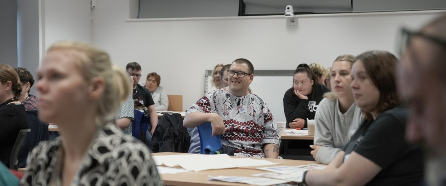 A group of people are sat at desks and listening to a talk.