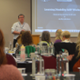 A room of people is listening to a presentation which is being delivered at the front.