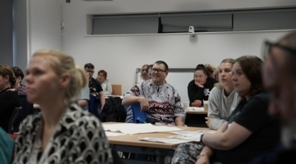 A group of people are sat at tables and listening to a presenter.