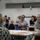 At the centre of a room of people, a man is smiling as he watches a presentation.